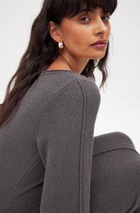 Vêtement en maille grise à manches longues avec un tissu texturé. Il présente un col rond et des détails de couture visibles sur les épaules et les manches.
