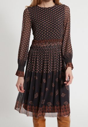 Kobieta w czarnej, przezroczystej sukience z długim rękawem, z pomarańczowymi okrągłymi i liściastymi wzorami, marszczoną talią i brązowymi kozakami za kolano.