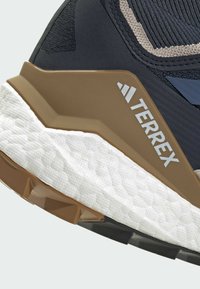 Terrex Wanderschuh mit texturiertem schwarzen Obermaterial, beigen Akzenten, brauner Zwischensohle und weißer gepolsterter Außensohle mit schwarzem Profil.