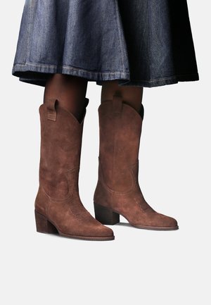 Bottes mi-mollet en daim marron avec des talons blocs bas portées avec des collants foncés et une jupe en jean bleu sur un fond uni.