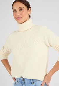 Jersey de cuello alto en crema, suave y con textura, con un yugo estampado y mangas ajustadas. Combinado con unos jeans de color azul claro.