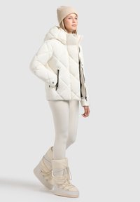 Chaqueta acolchada blanca tipo plumón con capucha, cremalleras frontales y puños elásticos, combinada con leggings crema y botas de nieve beige.
