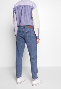 Conjunto masculino que incluye una camisa de botones multicolor con rayas moradas, azules y amarillas, combinada con jeans de mezclilla azul claro y zapatillas blancas.