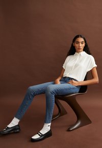 Witte blouse met korte mouwen, blauw slim-fit jeans, zwarte loafers met witte accenten, zittend op een bruine, gebogen stoel tegen een bruine achtergrond.