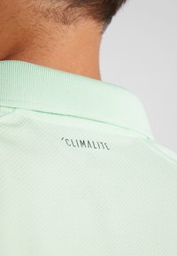 Polo vert clair en tissu texturé, avec un col strié et un logo "Climalite" noir au dos.