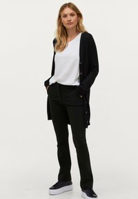 Sort cardigan med knapper, sammen med en hvid bluse og sorte bukser. Modellen har sorte sneakers på og står med hænderne i lommerne.