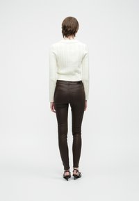 Osoba stojaca chrbtom, oblečená v bielom pletenom svetri, hnedých úzkych nohaviciach, čiernych podpätkoch a s viditeľným tetovaním na krku.