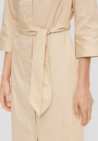 Beiges, tailliertes Kleid mit Dreiviertelärmeln, glatter Textur und gerader Silhouette; verfügt über einen gebundenen Stoffgürtel in der Taille.