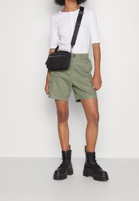 Pantaloni cargo verde oliva con cinque tasche, abbinati a un top bianco aderente a maniche lunghe e stivali da combattimento neri con lacci. Borsa a tracolla nera.