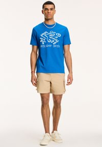 Tricou din bumbac albastru cu text grafic alb "VACANȚE OFTEN," asortat cu pantaloni scurți bej din bumbac. Include guler rotund și adidași masivi.