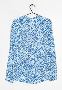 Blusa azzurra a maniche lunghe con motivo floreale bianco, orlo arrotondato e polsini con bottoni. Il tessuto appare leggero e ha una superficie liscia. Appesa a un gancio.