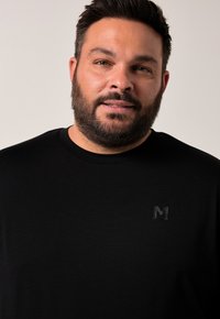 Svart kortärmad t-shirt tillverkad av mjukt tyg med en diskret präglad "M"-logotyp på bröstet. Enkel, klassisk design med rund halsringning.