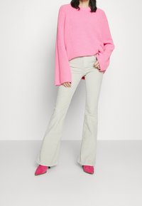 Rosa Strickpullover mit weiten Ärmeln, kombiniert mit beigen Schlaghosen und rosa Stiefeletten. Der Pullover hat ein strukturiertes Muster und eine lockere Passform.