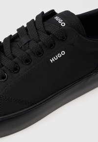 Crne niske tenisice s teksturiranim materijalom, debelim crnim vezicama i bijelim "HUGO" brendiranjem sa strane i na jeziku.