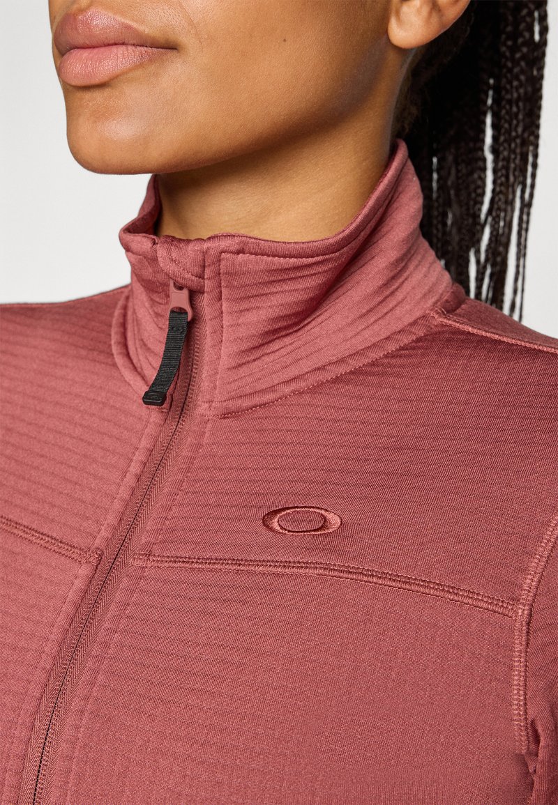 Een close-up van de nek en kin van een persoon die een roze jacket met rits en hoge kraag draagt, voorzien van een logo, waarbij de textuur van de stof goed in beeld komt.