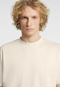 Helebeežika puuvillane t-särk ümmarguse kaelusega, lühikeste varrukatega ja vasakul rinnal peenelt trükitud logoga. Sile tekstuur.