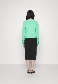 Blusa verde brilhante com bolinhas e mangas flare, combinada com uma saia preta até ao joelho e sapatos de salto pretos com tiras.