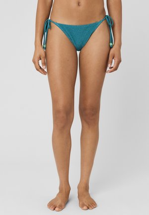 Femme portant un bas de bikini bleu sarcelle avec des liens latéraux et des perles, debout pieds nus devant un fond gris uni.