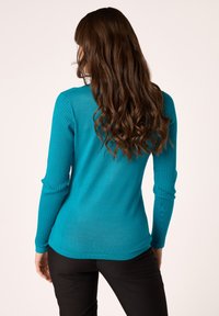 Maglione lungo a maniche lunghe color turchese con design aderente e texture morbida, caratterizzato da un orlo leggermente curvo e polsini accentuati.