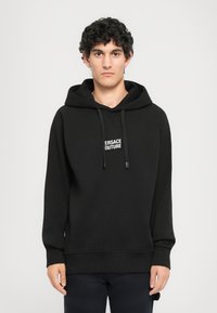 Čierna mikina s kapucňou a logom "VERSACE COUTURE." Obsahuje rebrované manžety a lem, vyrobená z jemnej, hrubej tkaniny.