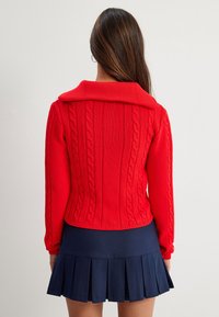 Czerwony sweter w warkoczowy wzór z szerokim kołnierzem, posiadający ściągacze przy mankietach i u dołu. Nosi się go na granatowej plisowanej spódnicy, podkreślającej dopasowany krój.