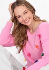 Rózsaszín V-kivágású pulóver, színes szalagokkal díszítve narancssárga, zöld, piros és kék színekben. A modellen világoskék farmernadrág van.