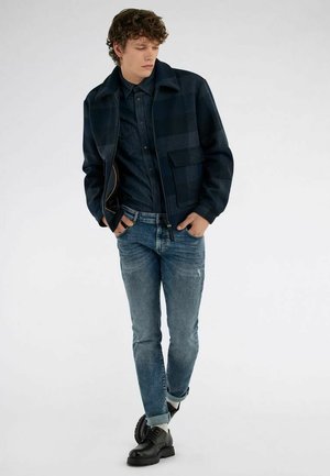 Giovane uomo con capelli ricci che indossa una giacca a quadri scura, una camicia scura, jeans blu risvoltati, calzini bianchi e scarpe nere stringate, in piedi con le mani nelle tasche.