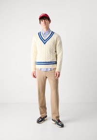 Maglione di cotone a maglia crema con strisce blu, indossato sopra una camicia colletto azzurro chiaro. I pantaloni beige e le sneakers multicolori completano il look.