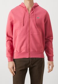 Felpa con cappuccio rosa con zip, dotata di cappuccio e tasche frontali, con un piccolo logo verde ricamato sul petto. Realizzata in tessuto morbido e strutturato.