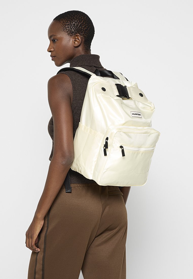 Mochila hecha de material blanco brillante, con dos bolsillos frontales con cremallera, una tapa superior y correas ajustables negras. Etiqueta de marca notable visible.