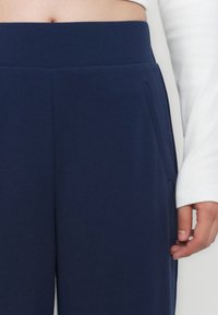 Navyblauwe strakke broek van een gladde stof, met een brede tailleband en zijzakken. Er ligt een wit langemouwtopje in de buurt.