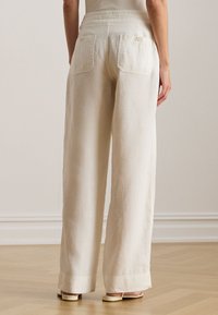 Plati siaurė, šviesios smėlio spalvos lininės kelnės su aukštu liemeniu ir dviem užsegamais kišenėmis gale. Lygios tekstūros ir laisvo kirpimo.