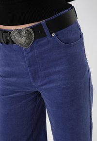 Pantalones de pana azul con una textura acanalada, que cuentan con un cinturón negro y una hebilla decorativa en forma de corazón de plata.