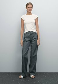Witte fitted t-shirt gecombineerd met oversized donkergrijze jeans met een rechte snit. Het model draagt witte sandalen. Neutrale achtergrond.