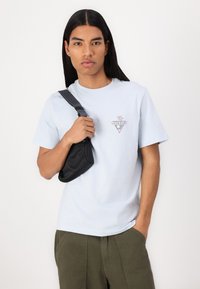 T-shirt en coton bleu clair avec un logo en triangle présentant un design floral. Le modèle tient un petit sac noir avec une sangle. Un pantalon vert est visible.