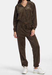 Completo in velluto marrone con felpa zip e pantaloni coordinati. La felpa presenta una tasca a marsupio e polsini in costina. I pantaloni hanno una vita elastica con cordoncino.