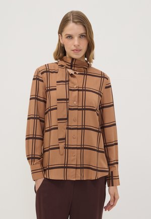 Mujer que lleva una blusa marrón de cuadros con botones y un lazo en el cuello, combinada con pantalones marrón oscuro, de pie frente a un fondo liso.