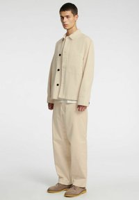 Beżowa bawełniana kurtka z czarnymi guzikami i kieszenią na piersi, zestawiona z luźnymi beżowymi spodniami i jasnobrązowymi butami.