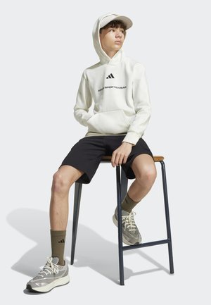 Sudadera blanca con una marca negra, pantalones cortos negros, zapatillas beige y calcetines verde oliva. Sentado en un taburete de bar, fondo de luz natural.