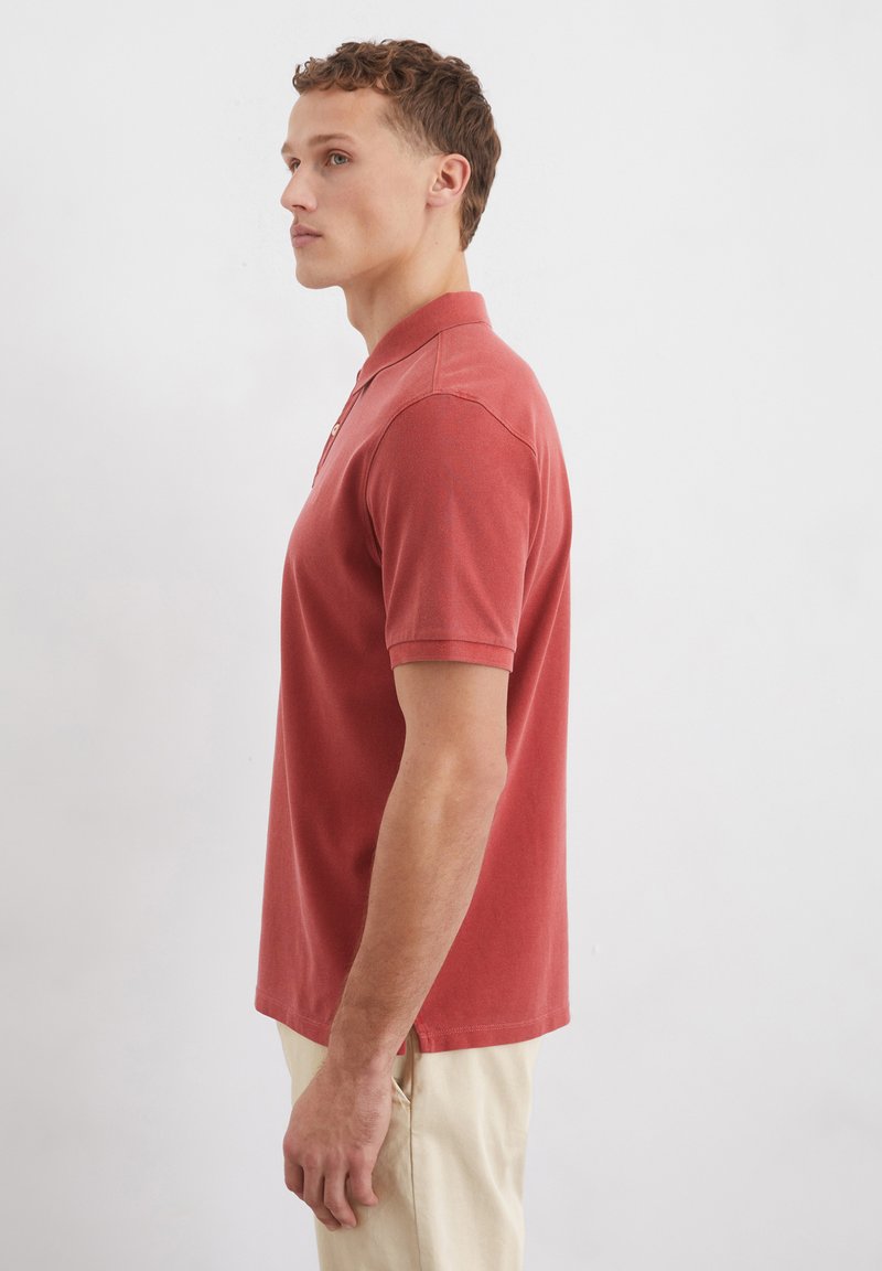 Rotes Poloshirt mit kurzen Ärmeln, Kragen und subtiler Textur. Getragen mit hellen Hosen, die ein Seitenprofil des Models zeigen.