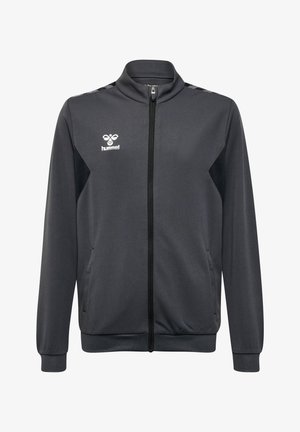 Dreng med blonde dreadlocks iført en sort Hummel zip-op sportsjakke, hænderne i lommerne, ser mod højre på en ensfarvet baggrund.