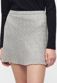 Femme portant une jupe courte en maille torsadée gris clair et un haut noir à manches longues, les mains reposant de chaque côté.