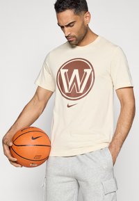 Krémové bavlnené tričko s hnedým kruhovým logom so "W" v strede, pričom je kombinované so šedými teplákmi a drží oranžový basketbal.