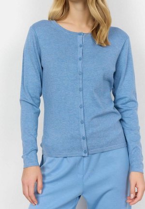 Femme portant un cardigan bleu clair boutonné avec des manches longues et un pantalon ample assorti, les mains détendues le long du corps.