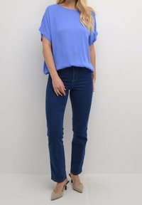 Blaues kurzärmeliges Oberteil kombiniert mit dunkelblauen Skinny Jeans und beigen Absatzmules. Glatter Stoff, lässige Passform und schlichtes Design.