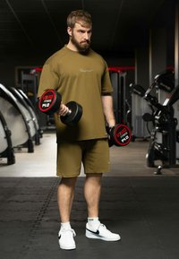 Maglietta sportiva verde oliva e pantaloni corti. Tiene dei manubri neri con etichette rosse. Scarpe da ginnastica bianche. Ambiente da palestra con attrezzature sullo sfondo.