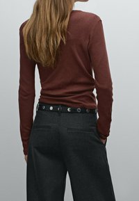 Bruine langemouwen shirt gecombineerd met een grijze broek. De outfit bevat een zwarte riem met zilveren ronde accenten, die de taille accentueert.