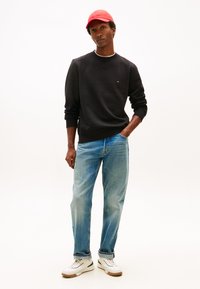 Fiatal férfi piros sapkában, fekete pulóverben, világoskék farmerral és fehér tornacipőben, egyik kezét zsebében tartva, fehér háttér előtt áll.