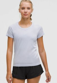 Jonge vrouw met blond haar in een staart, gekleed in een wit sportshirt met korte mouwen en zwarten hardloopshorts, leunend tegen een effen achtergrond.
