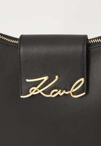 Svart lærveske med et gulltonet "Karl"-logo på låsen, med en glatt tekstur og glidelås på toppen.